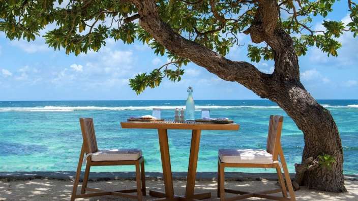 Ein gedeckter Tisch mit zwei Stühlen steht unter einem Baum am Strand mit Blick auf das Meer.