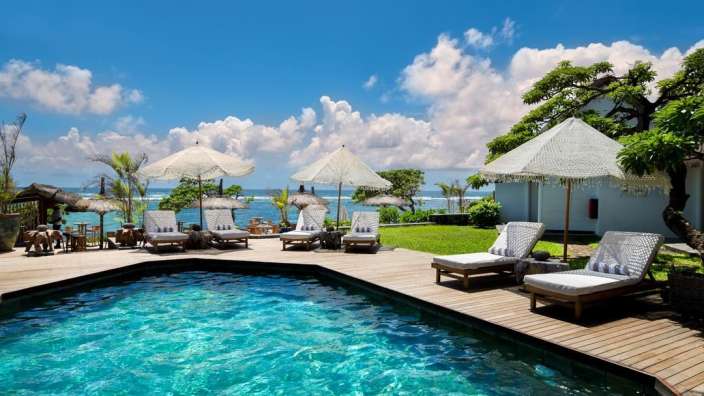 Ein luxuriöser Pool mit Liegestühlen und Sonnenschirmen steht am Meer unter einem blauen Himmel.