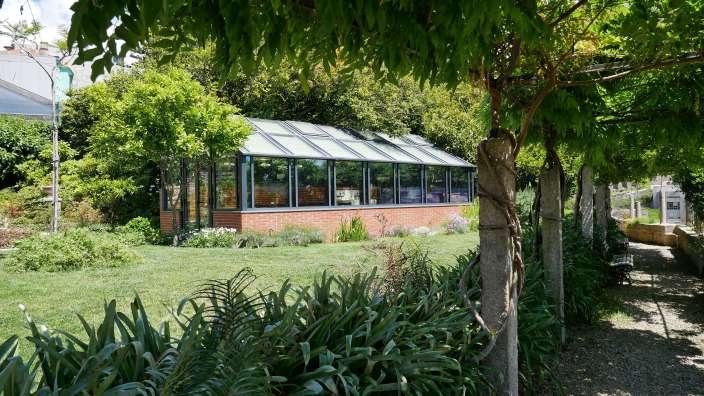 Ein Glashaus steht auf einem gepflegten Rasen, umgeben von üppigem Grün und einem überdachten Weg mit Sitzbänken.