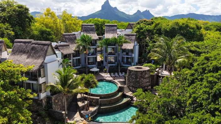 Eine Drohnenaufnahme zeigt eine Hotelanlage mit Pools und strohgedeckten Gebäuden im Vordergrund, umgeben von üppiger Vegetation und mit einem zerklüfteten Bergmassiv im Hintergrund.