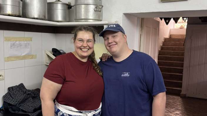 Ein Mann und eine Frau stehen in einer Küche und lächeln in die Kamera, beide tragen Schürzen mit einem blauen Muster.