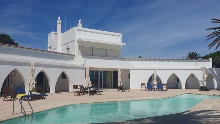 Ein weißes Gebäude mit mehreren Bögen und einem Swimmingpool im Vordergrund steht unter einem blauen Himmel.