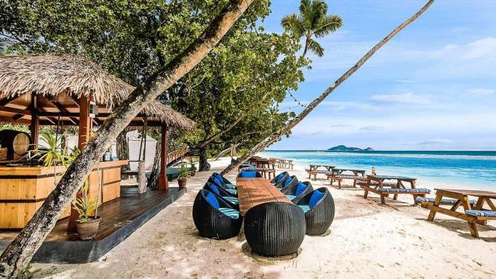 Ein Strand mit einer Bar und Tischen im Schatten von Bäumen am Meer.