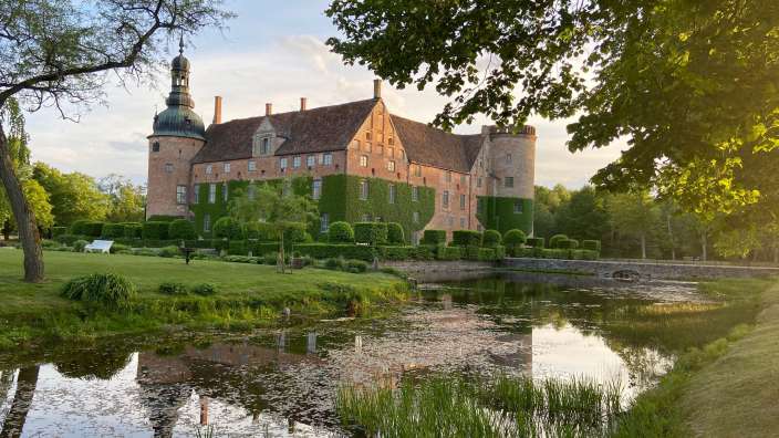 Ein Schloss mit einem Turm ist von einem Teich und Grünflächen umgeben, mit Bäumen im Vordergrund, die das Sonnenlicht reflektieren.