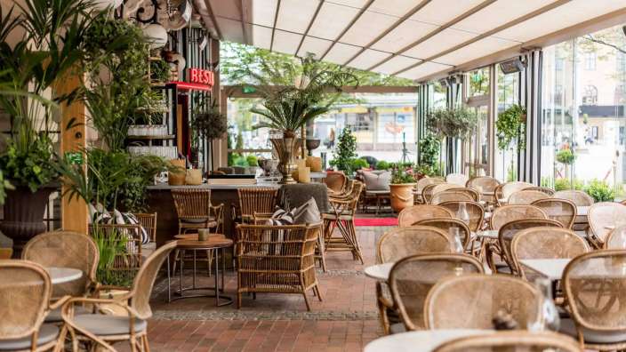 Ein offenes Restaurant mit Rattanmöbeln, Pflanzen und einer Markise ist bei Tageslicht zu sehen.