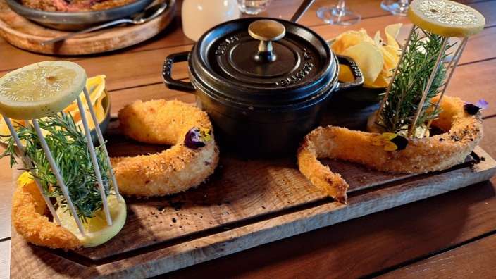 Die Nahaufnahme zeigt ein rustikales Holzbrett mit zwei frittierten Gerichten, Garnelen und einer Mini-Schmortopf, serviert mit Garnitur und Chips.