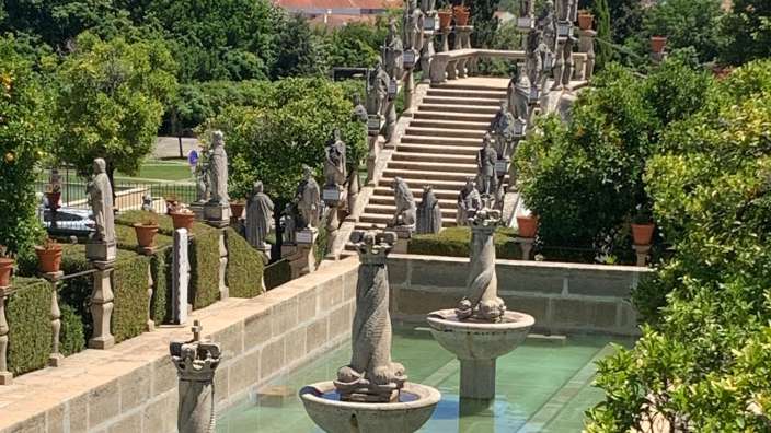 Ein Garten mit symmetrischen Skulpturen, einer Steintreppe und einem Brunnen mit türkisfarbenem Wasser.