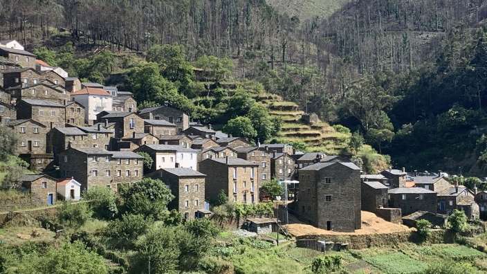 Eine kleine Bergdorf-Siedlung mit Steinhäusern und terrassierten Feldern ist vor einem bewaldeten Hang zu sehen.