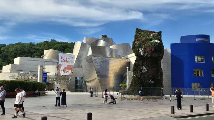 Das Bild zeigt die skulpturale Gestaltung des Guggenheim Museums Bilbao, mit dem Blumen-Welpen als Blickfang und dem blauen Gebäude im Hintergrund.