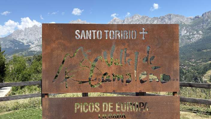 Ein rostfarbenes Schild mit ausgeschnittenen Buchstaben, das "SANTO TORIBIO" und "PICOS DE EUROPA" anzeigt, steht vor einer Bergkulisse und einem blauen Himmel mit Wolken.