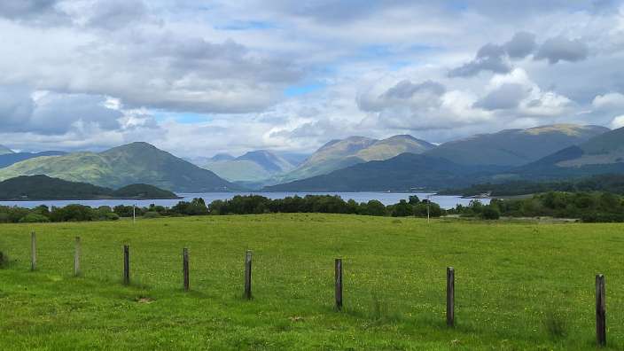 Grünes Feld mit Holzzaun vor einer Bucht mit Bergen im Hintergrund unter bewölktem Himmel.