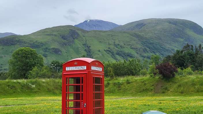 Ein rotes Telefonhäuschen steht auf einer grünen Wiese mit gelben Blumen, vor einer bergigen Landschaft mit einem schneebedeckten Gipfel.