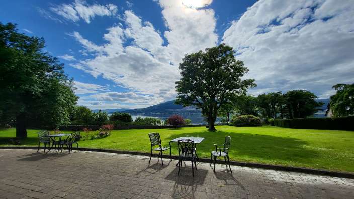Ein Tisch mit zwei Stühlen steht auf einer gepflasterten Terrasse vor einer grünen Rasenfläche mit Bäumen und einem See im Hintergrund.