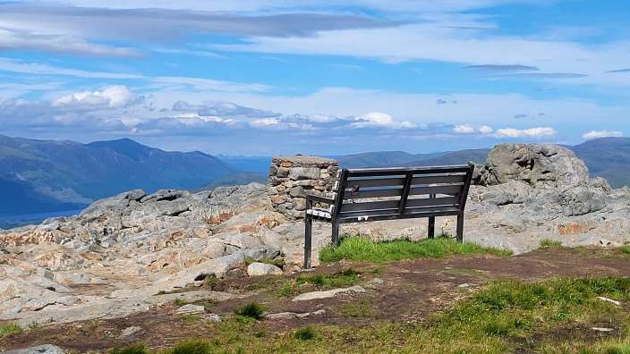 Ein hölzerner Sitzplatz steht auf einem felsigen Berggipfel mit Blick auf einen See und Berge unter einem blauen Himmel mit Wolken.