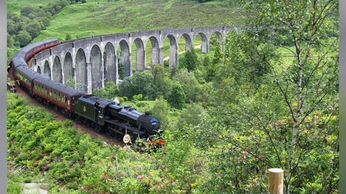 Eine Dampflokomotive mit roten Waggons fährt über eine hohe Steinbogenbrücke, umgeben von üppigem Grün.