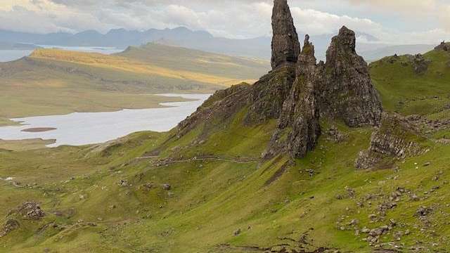 Die dramatischen Felsformationen des Old Man of Storr ragen über eine grüne, hügelige Landschaft mit einem See im Hintergrund auf.