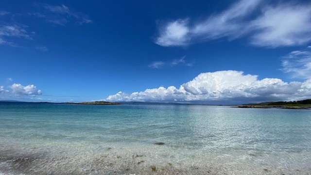 Ein ruhiger Strand mit klarem, türkisfarbenem Wasser und einer weiten, blauen Himmelkulisse mit dramatischen Wolken.