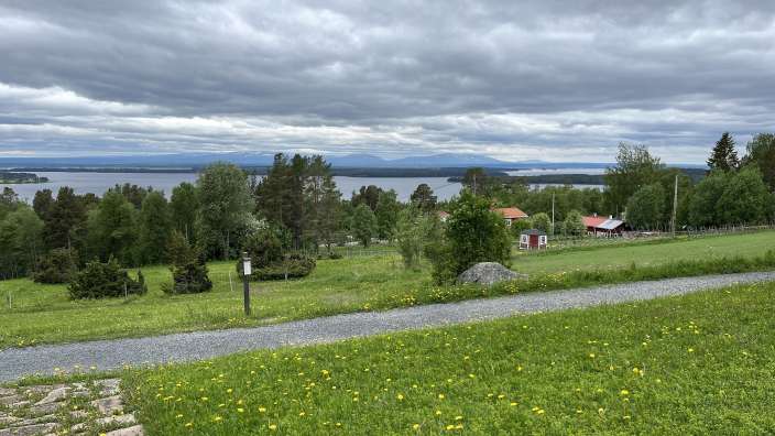 Eine idyllische Landschaft mit einem See, Bergen im Hintergrund und einem grasbewachsenen Hang, auf dem kleine Häuser und Bäume stehen, unter einem dramatischen Himmel mit Wolken.