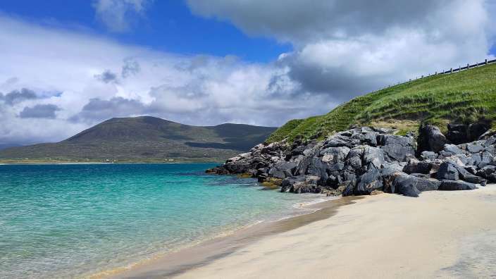 Das Bild zeigt eine felsige Küste mit einem türkisfarbenen Meer, einem sandigen Strand und bewachsenen Hügeln im Hintergrund unter einem bewölkten Himmel.