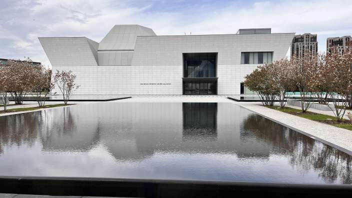 Das Bild zeigt das Aga Khan Museum mit einem großen, spiegelnden Wasserbecken im Vordergrund und Bäumen mit blühenden Zweigen an den Seiten.