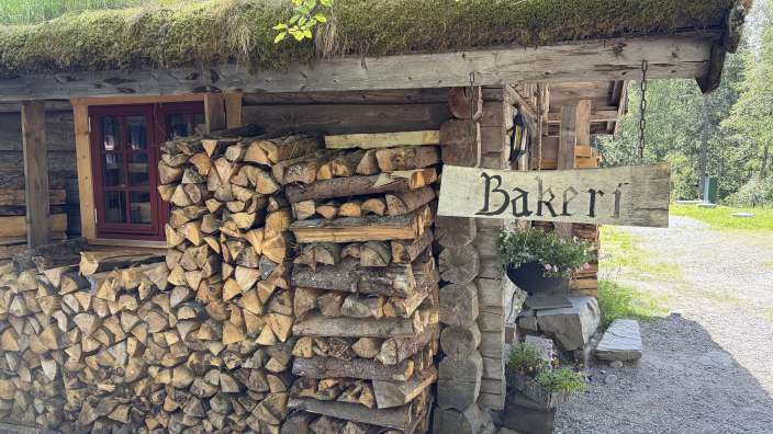 Ein Holzhaus mit Grasdach und einem Schild "Bäckerei" steht vor einem Wald.