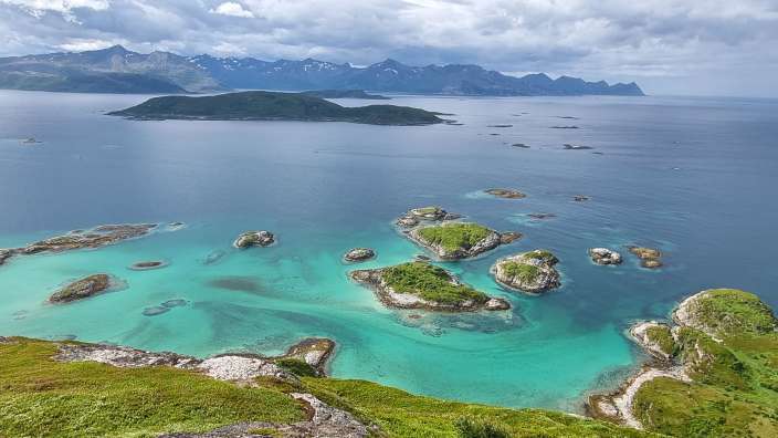 Eine von Inseln übersäte Küstenlandschaft mit türkisfarbenem Wasser und schneebedeckten Bergen im Hintergrund unter einem bewölkten Himmel.
