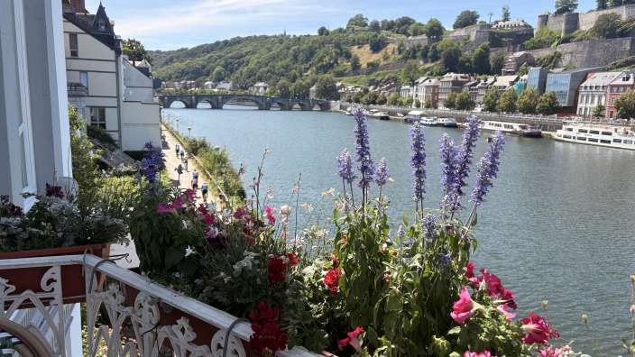 Ein Blick über einen Balkon mit Blumen auf einen Fluss mit Booten, einer Brücke und einer bergigen Stadtkulisse.