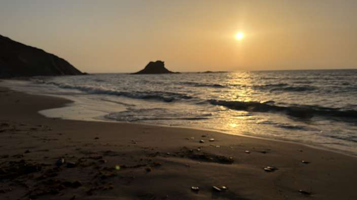 Ein ruhiger Strandblick bei Sonnenuntergang mit Wellen, die auf den Sand treffen, und einem kleinen Felsen im Meer.