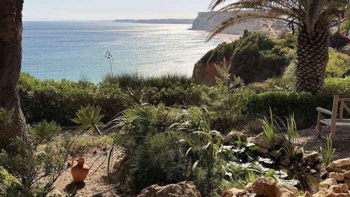 Das Bild zeigt eine mediterrane Küstenlandschaft mit einem Garten und dem Meer im Hintergrund.