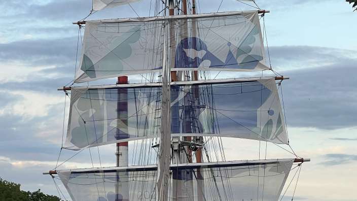 Ein großes Segelschiff mit mehreren weißen Segeln, auf denen blaue Muster und Text zu sehen sind, liegt in einem Kanal, gesäumt von Bäumen und Gebäuden.