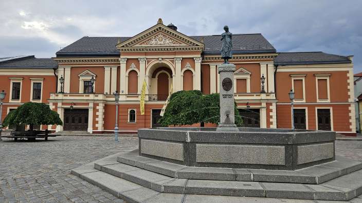 Ein Brunnen und eine Statue stehen auf einem gepflasterten Platz vor einem prächtigen orangefarbenen Gebäude mit einem verzierten Giebel und mehreren Fenstern.