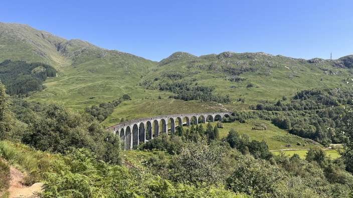 Ein eleganter Eisenbahnviadukt aus Stein mit vielen Bögen erstreckt sich über eine üppig grüne, hügelige Landschaft unter einem strahlend blauen Himmel.