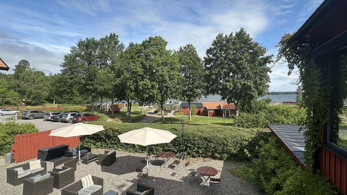 Eine Terrasse mit Sitzgelegenheiten und Sonnenschirmen ist mit Blick auf einen See von Bäumen und roten Holzhäusern umgeben.