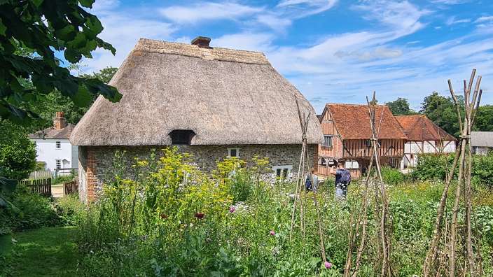 Ein Cottage mit Strohdach steht in einem üppigen Garten vor einer ländlichen Landschaft mit Fachwerkhäusern und einem blauen Himmel mit Wolken.