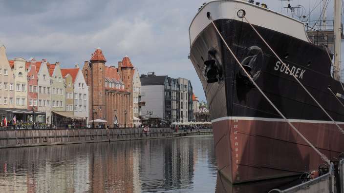 Das Bild zeigt ein Schiff namens "Sołdek", das in einem Hafen vor einer Reihe bunter Gebäude liegt.