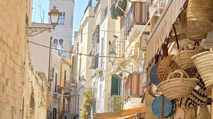 Menschen schlendern am Tag durch eine schmale Gasse in Bari, überragt von einem hohen Kirchturm und belebten Balkonen.