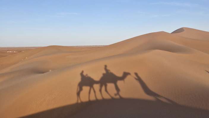 Schattenspiel eines Dromedar-Ausflugs in der marokkanischen Chigaga-Wüste bei tiefem Sonnenstand über Sanddünen.