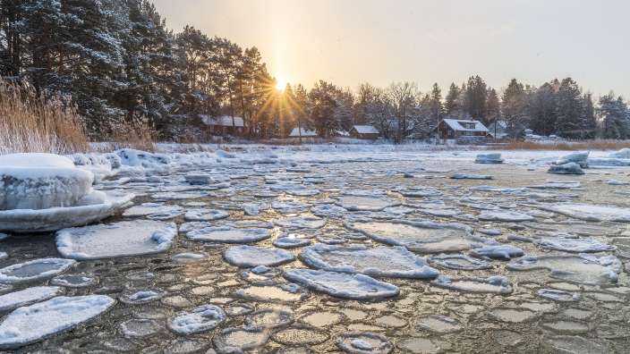 Wintersonne über dem eisbedeckten Meer vor dem estnischen Fischerdorf Altja mit verschneiten Holzhäusern und Bäumen.