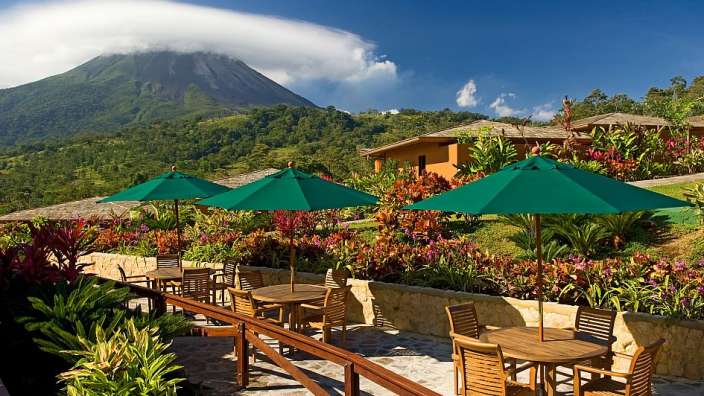Eine Terrasse mit Tisch und Stühlen und grünen Regenschirmen mit einem Vulkan im Hintergrund